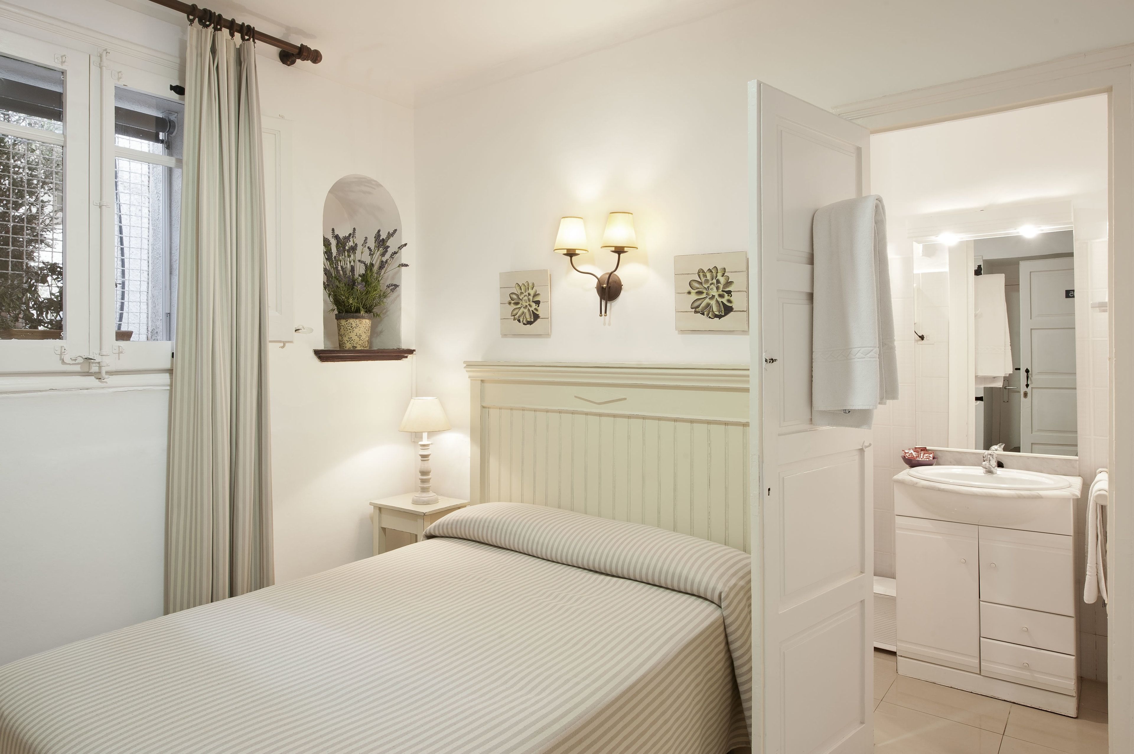 Een slaapkamer met een bed, nachtkastje, lamp, wanddecoratie en een raam met gordijnen.