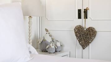 Un dormitorio con una cama blanca, una lámpara de noche y adornos en la pared, incluyendo una pieza de mimbre con forma de corazón y dos pequeñas figuras de pájaros.