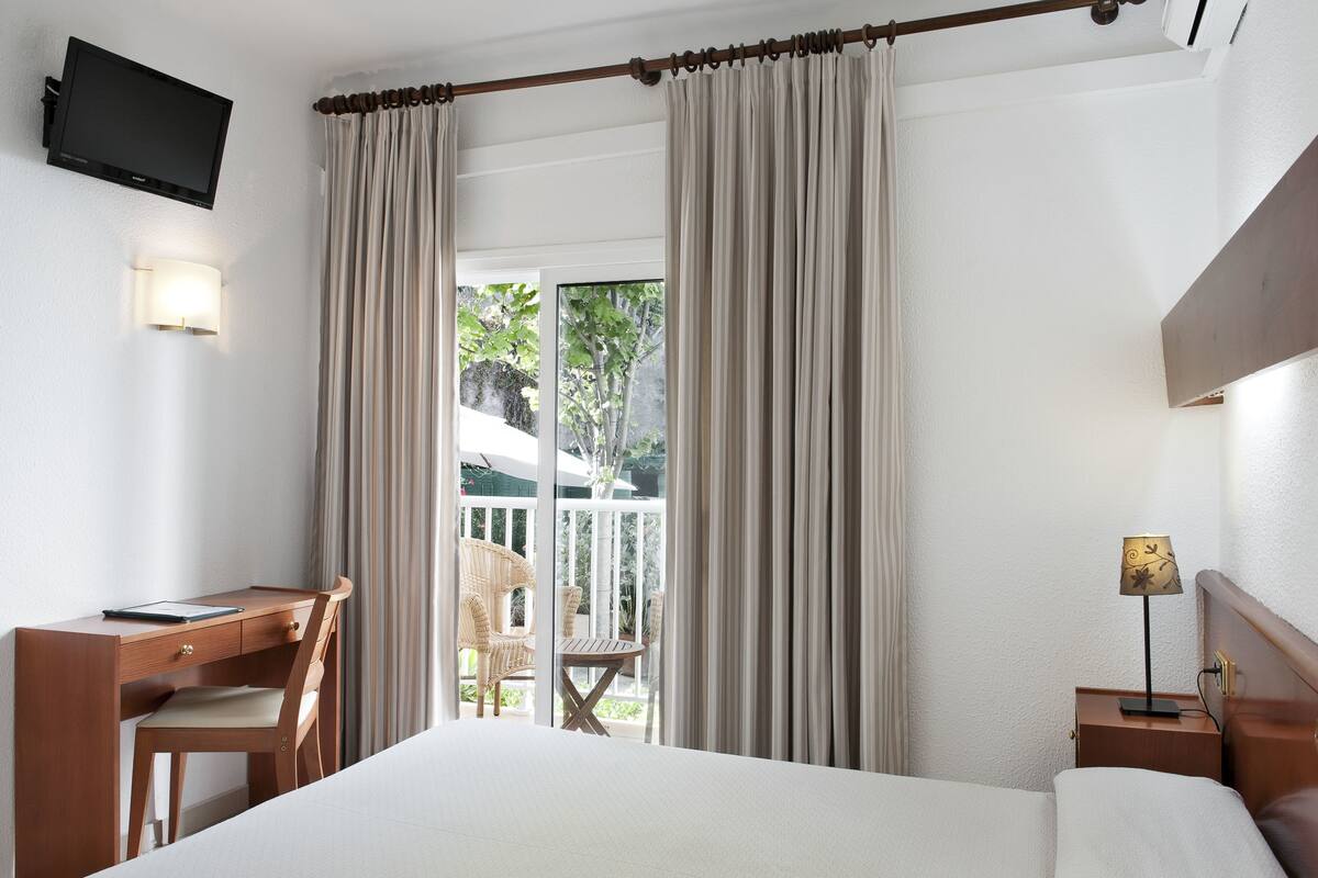 A hotel room with a bed, a desk with a chair, a small table, a TV mounted on the wall, and a balcony with curtains.
