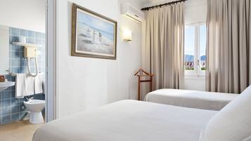 Habitación de hotel con dos camas, baño con pared de azulejos azules, un cuadro con escena de playa y vistas a las montañas desde la ventana.
