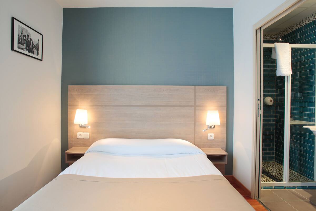 Ein Hotelzimmer mit einem Bett, zwei Nachttischlampen, einem Nachttisch und einem Duschbereich mit blauen Fliesen.