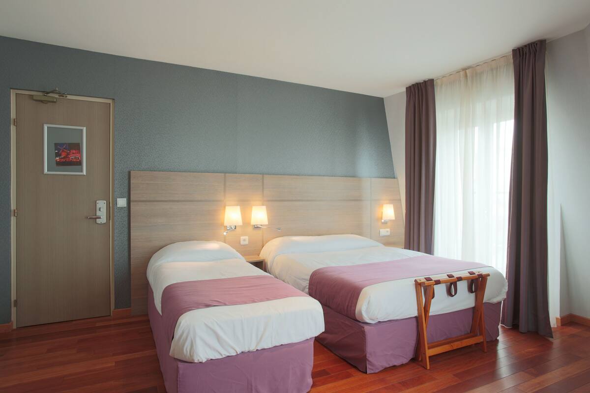 Ein Hotelzimmer mit zwei Betten, einem hölzernen Kopfteil, Nachttischlampen, einer Holzbank, einer Tür und einem gerahmten Bild an der Wand.
