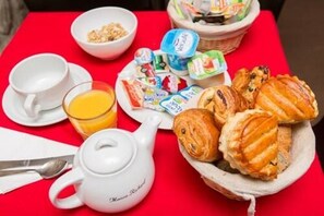 A breakfast setup with a teapot, cups, a glass of orange juice, a bowl of cereal, a plate of pastries, and a basket of yogurt containers. - Grand Hôtel du Loiret (Paris)