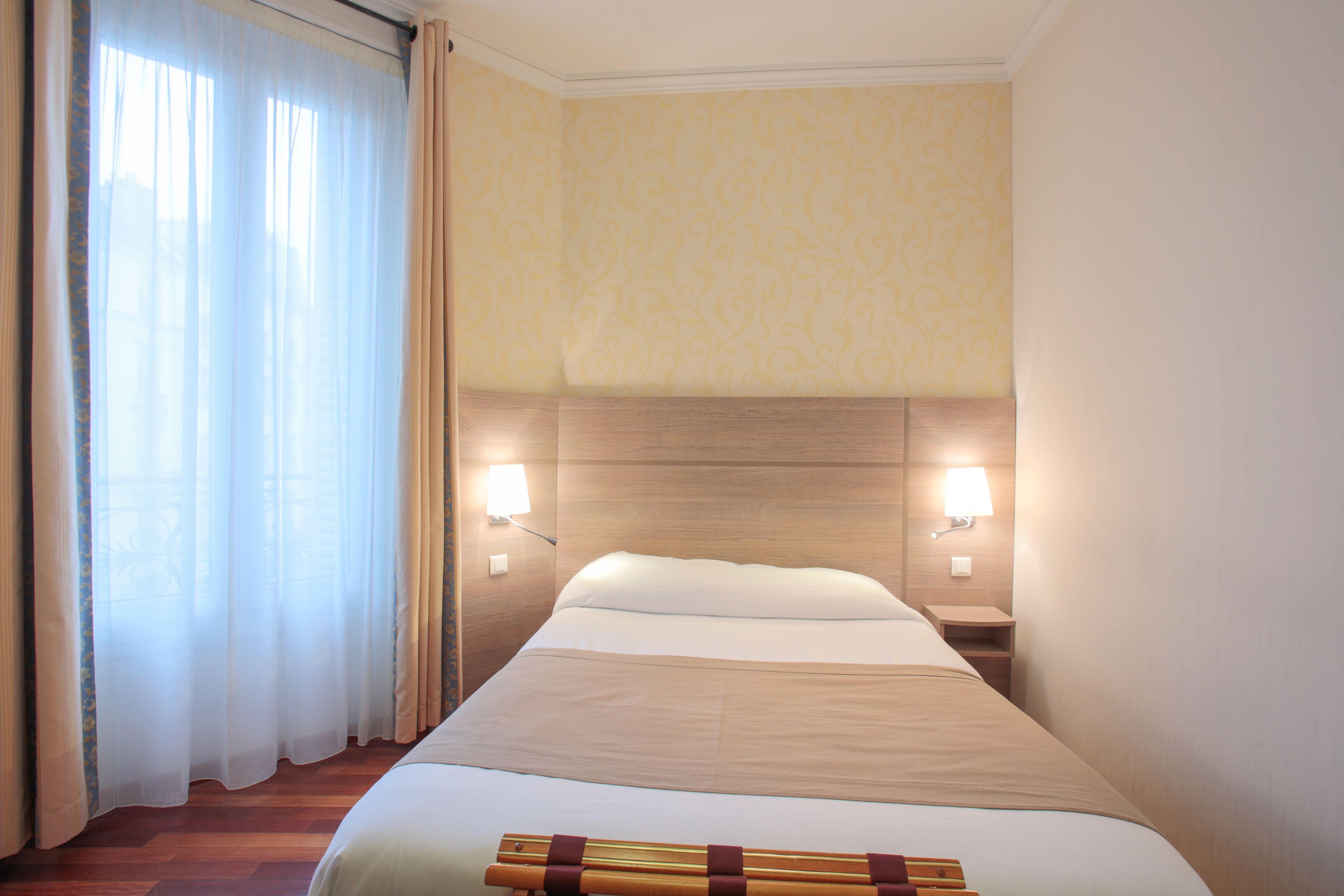 A hotel room with a bed, two bedside lamps, a nightstand, and a window with curtains.