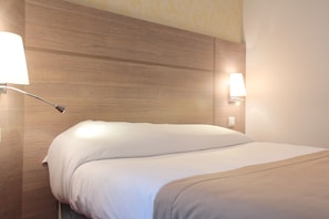 Cama bem arrumada com roupa de cama branca e bege, cabeceira de madeira e duas luminárias laterais fixadas na parede.