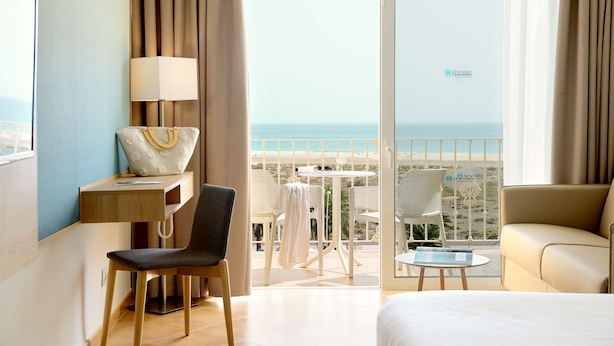 Habitación de hotel con cama, escritorio con lámpara, una silla, una mesita con una silla y un balcón con vista al mar.