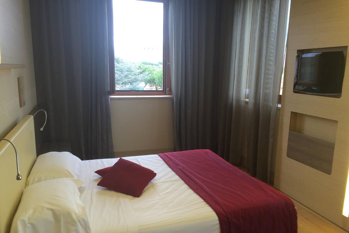Une chambre d’hôtel avec un lit, un téléviseur fixé au mur et une fenêtre avec des rideaux.