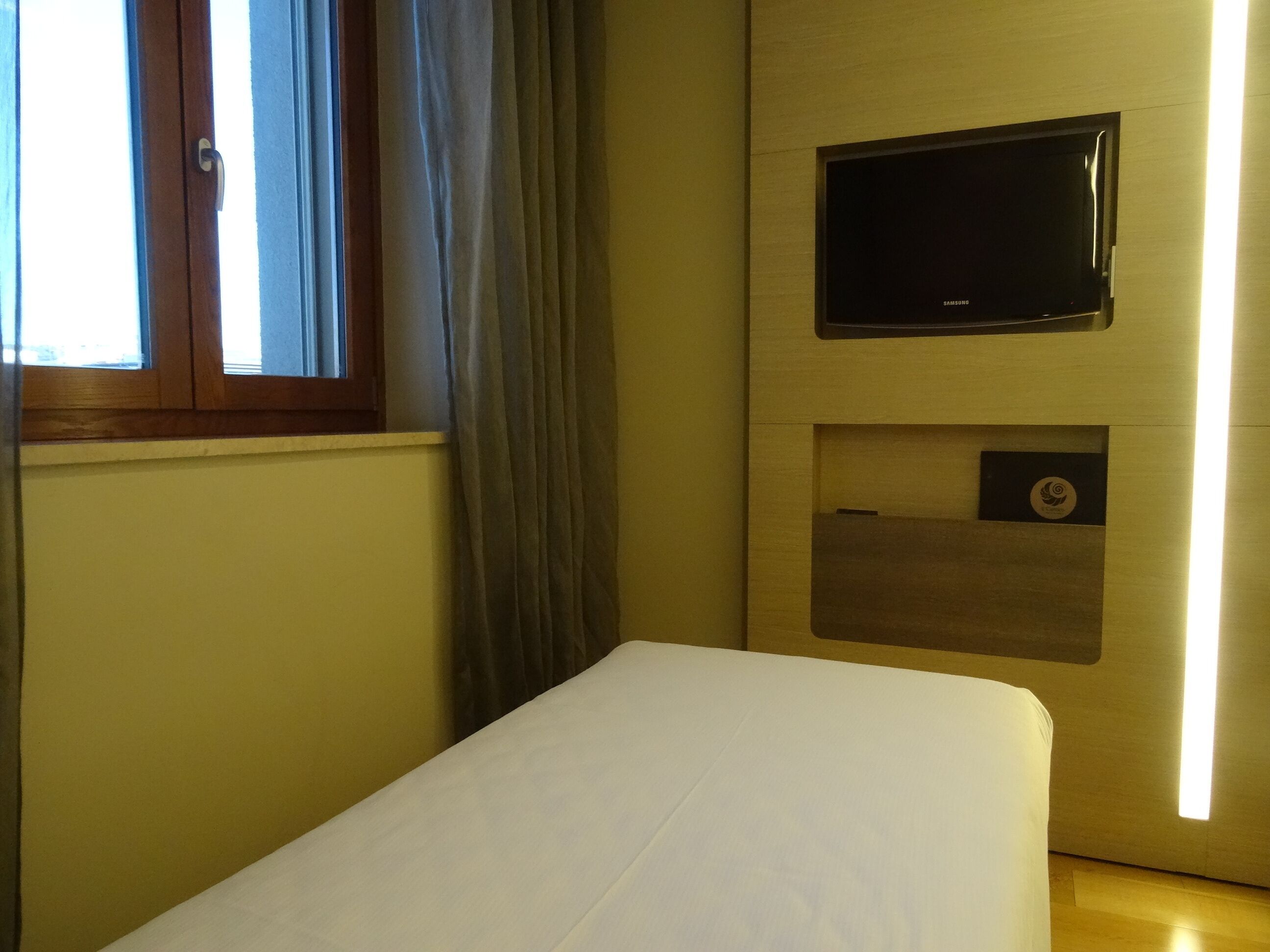 Una habitación de hotel con una cama, un televisor montado en la pared y una ventana con cortinas.