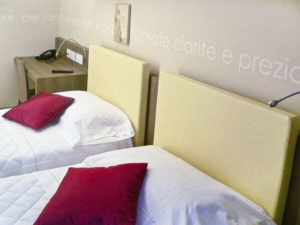 A hotel room with two beds, each with a white and red pillow. There is a nightstand with a phone and a lamp. The wall has a religious symbol and text. - Hotel Il Cantico St. Peter (Rome)