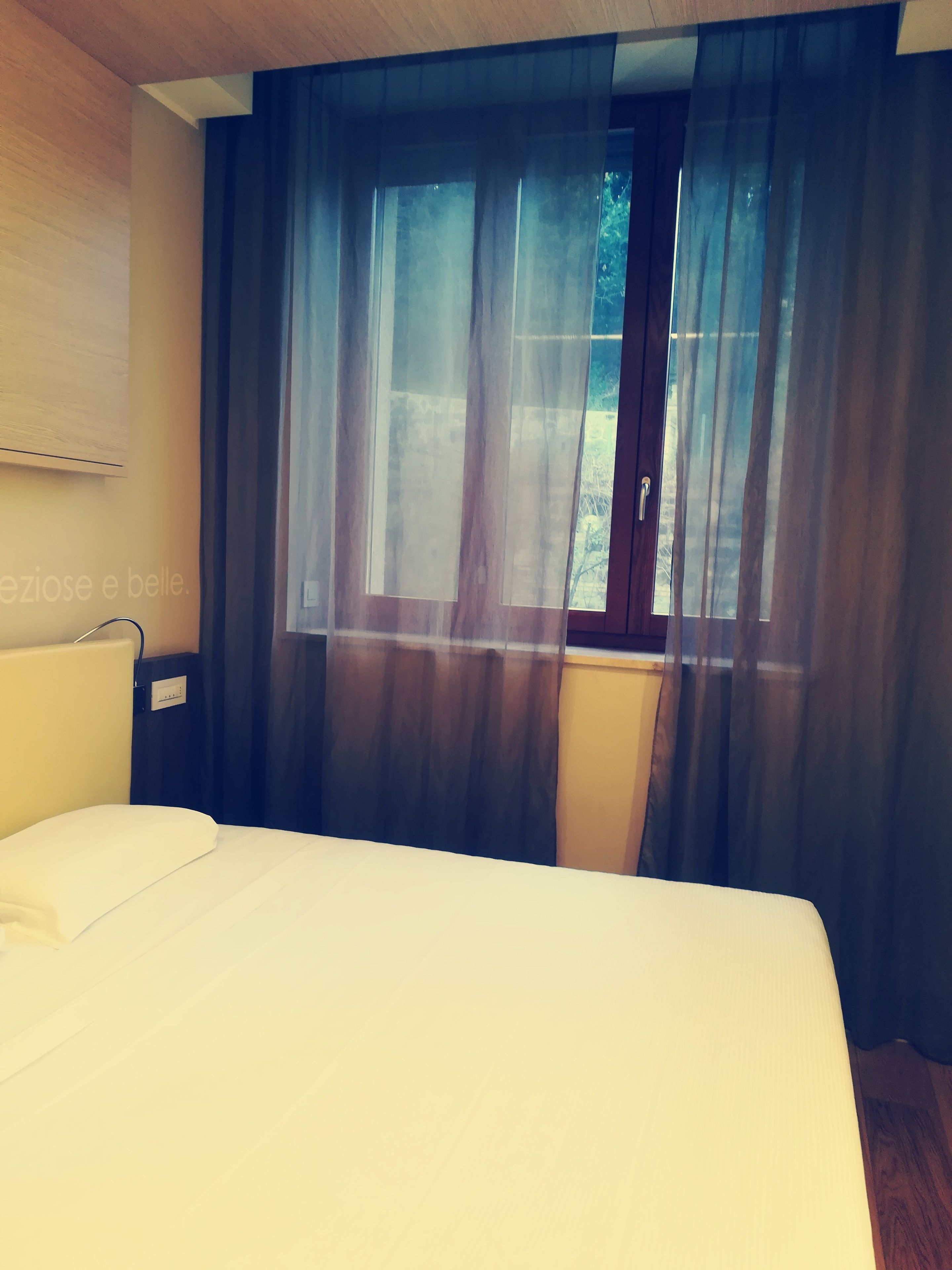 Une chambre d’hôtel avec un lit, une fenêtre avec des rideaux et un luminaire fixé au mur.