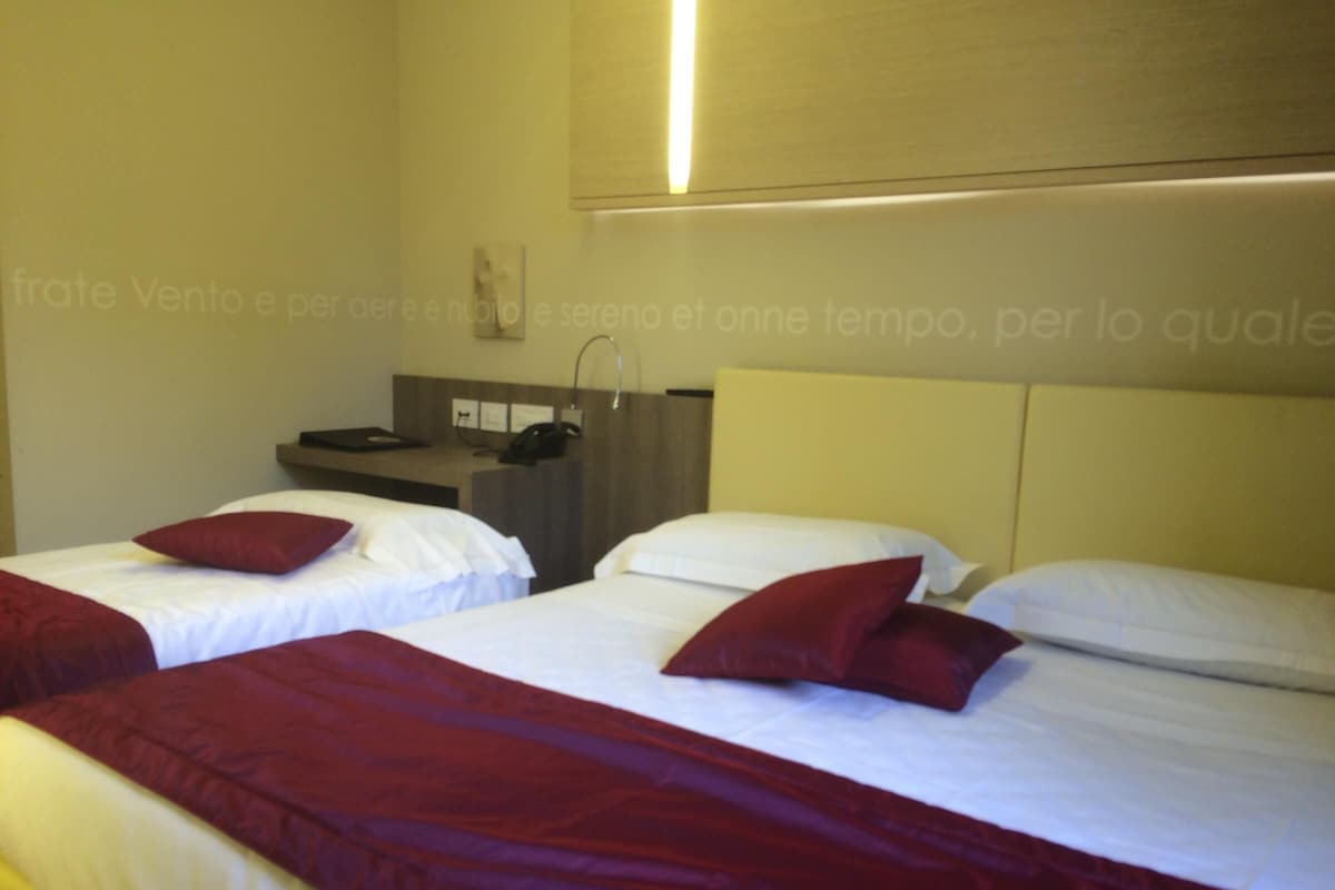 Une chambre d’hôtel avec deux lits, chacun étant équipé de draps blancs et d’oreillers rouges. On y trouve un bureau avec un téléphone et une petite étagère sur laquelle sont posés divers objets.