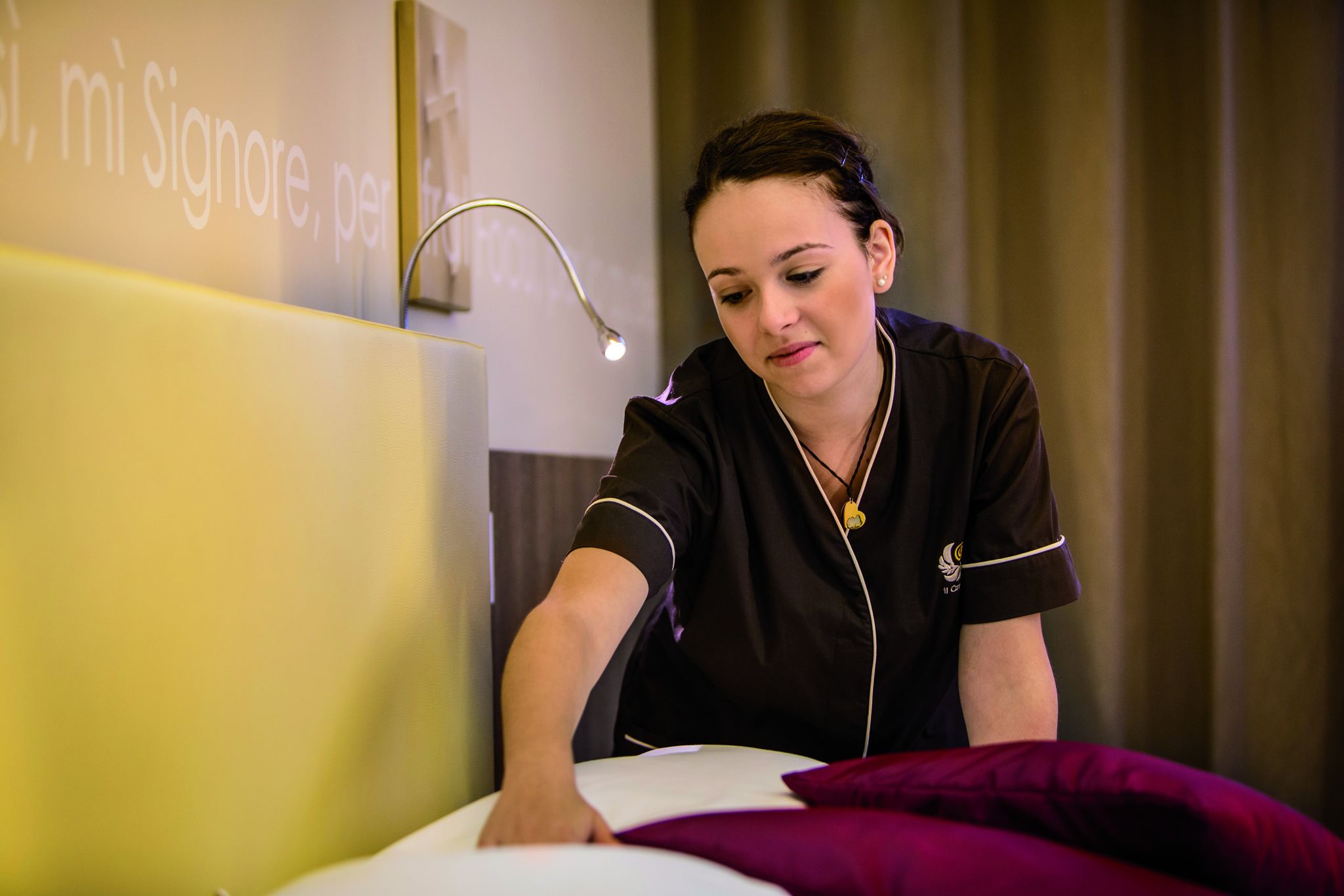 A person in a black uniform leaning over a bed with a white pillow and a purple blanket.