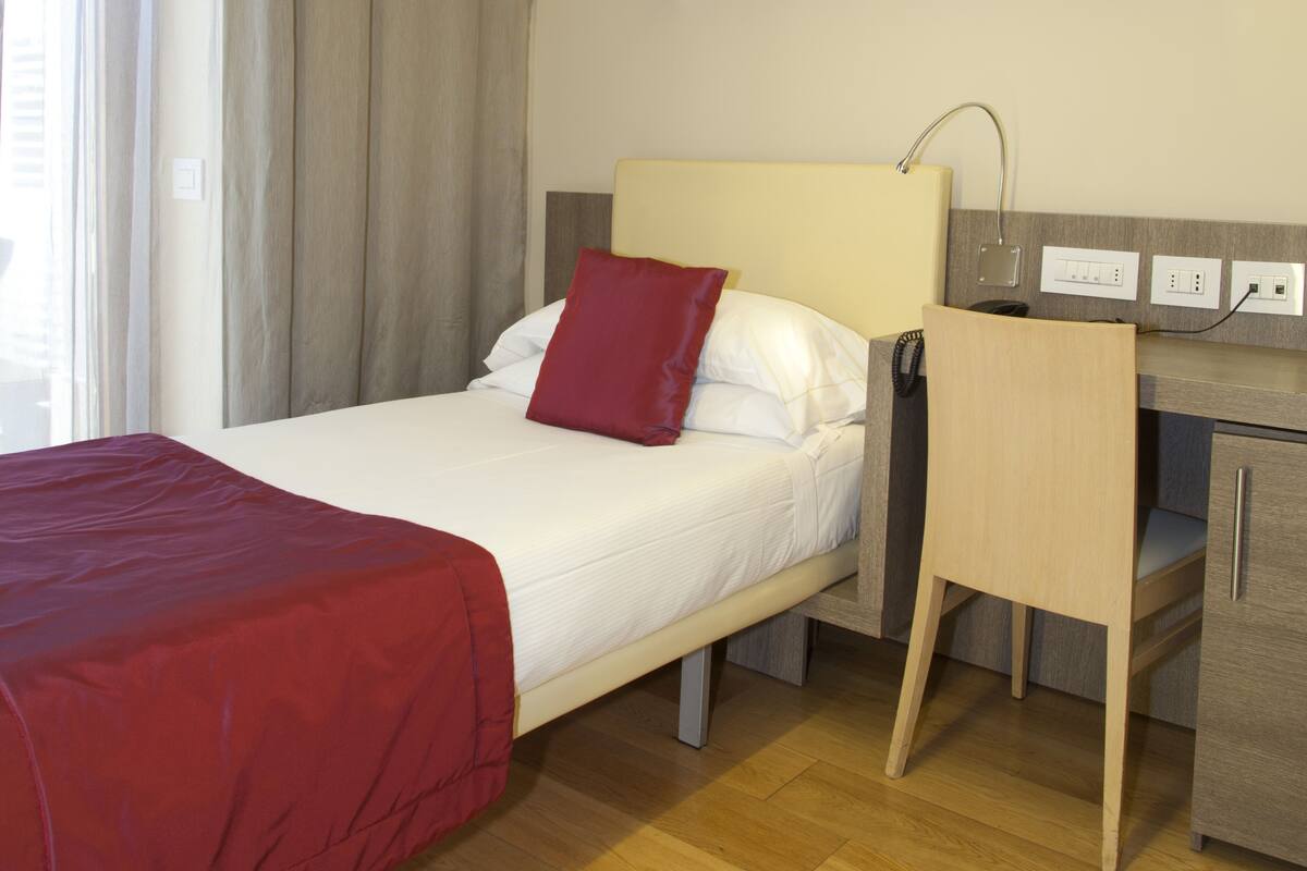 Une chambre d’hôtel avec un lit, un bureau et une chaise. Il y a une couverture rouge sur le lit et une lampe sur le bureau.