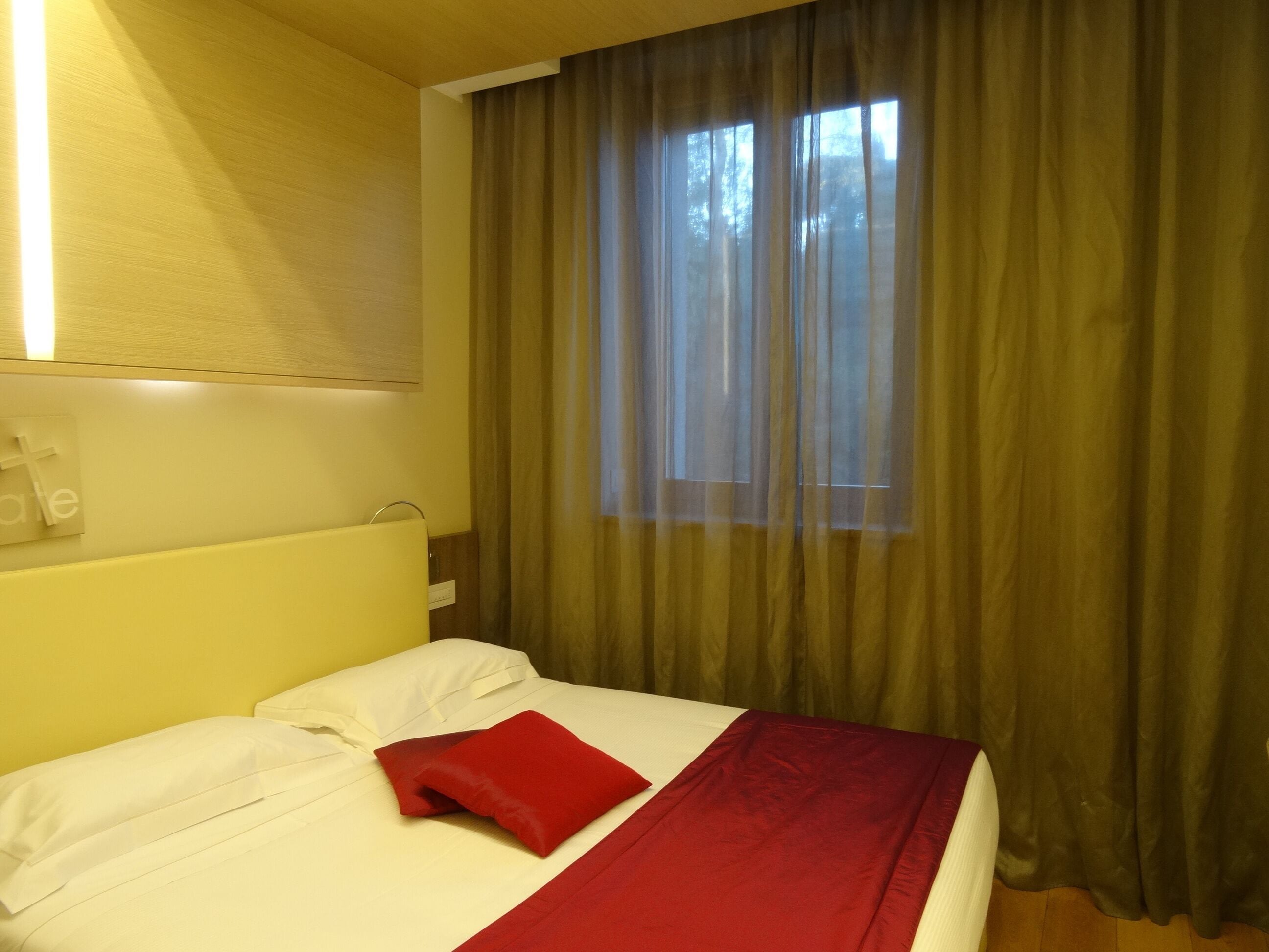Habitación de hotel con una cama, una ventana con cortinas, una lámpara en la pared y un cartel con una cruz.