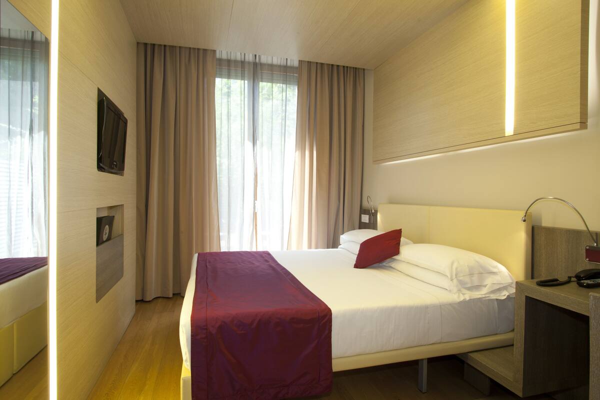 Une chambre d’hôtel avec un lit, une table de chevet, une lampe, une télévision et une fenêtre avec des rideaux.