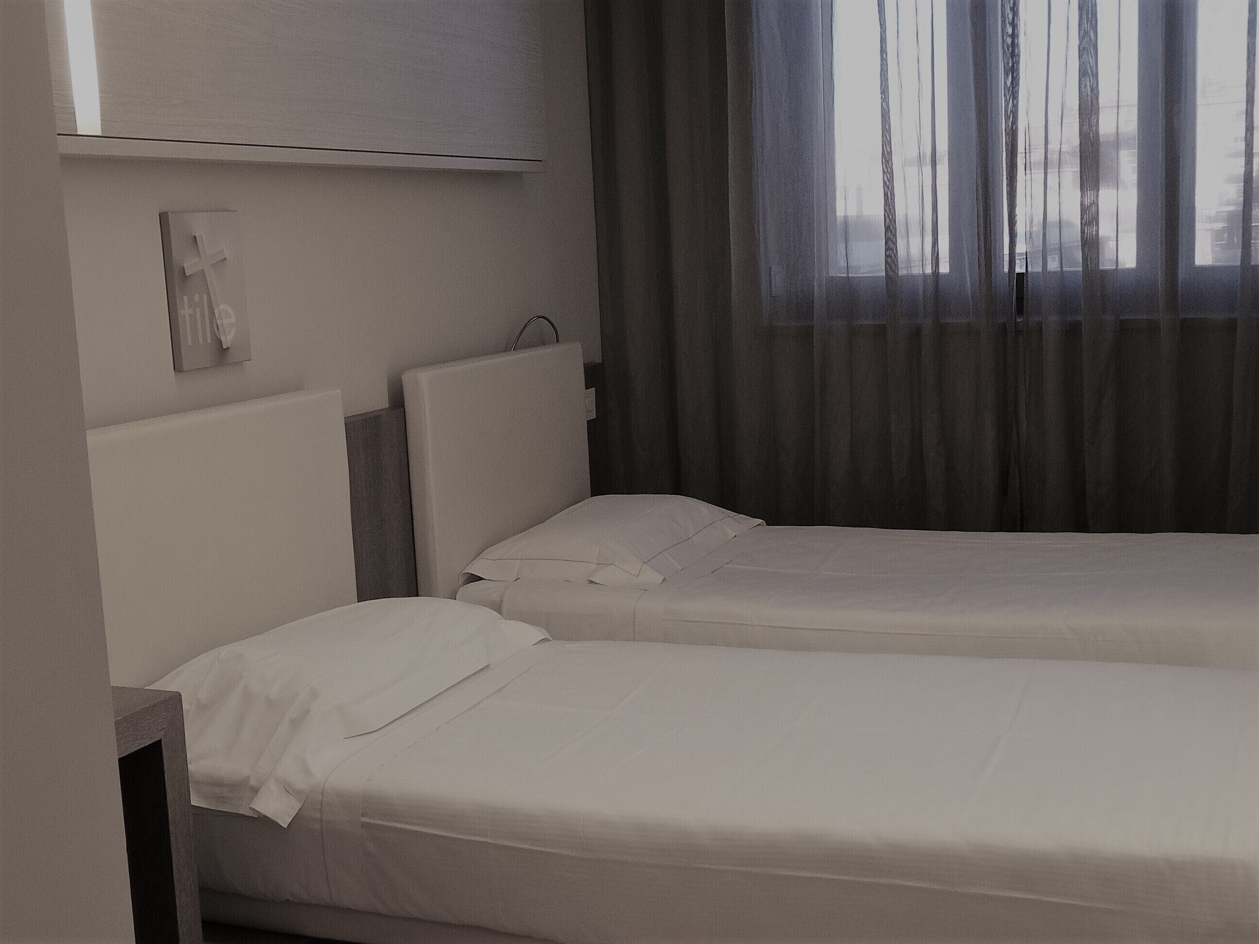 Una camera d'albergo con due letti singoli, un comodino, un telefono fissato al muro e una finestra con tende leggere.