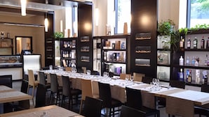 A modern restaurant with long communal tables, black chairs, and pendant lighting.