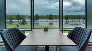 A room with a large table and two chairs, a view of a lake, and a cloudy sky. - Hayat Hospitality Oskemen (Oskemen)