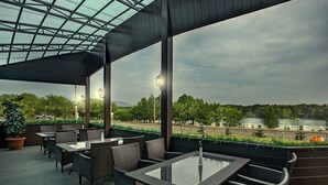 An outdoor seating area with wicker furniture and a glass table, overlooking a road and trees. - Hayat Hospitality Oskemen (Oskemen)