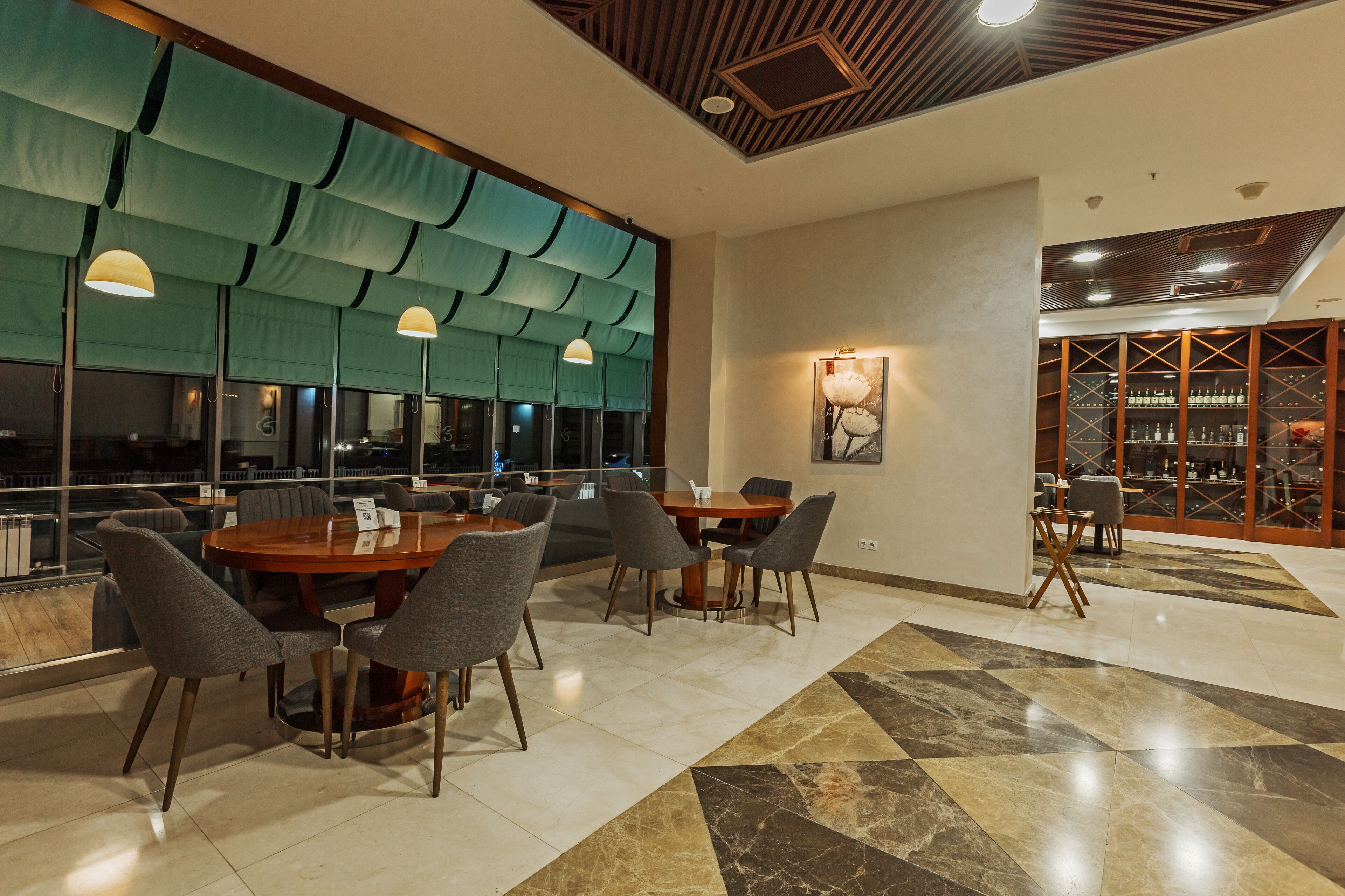 A modern dining area with a round table and chairs, a large window with green curtains, and a wine rack in the background.