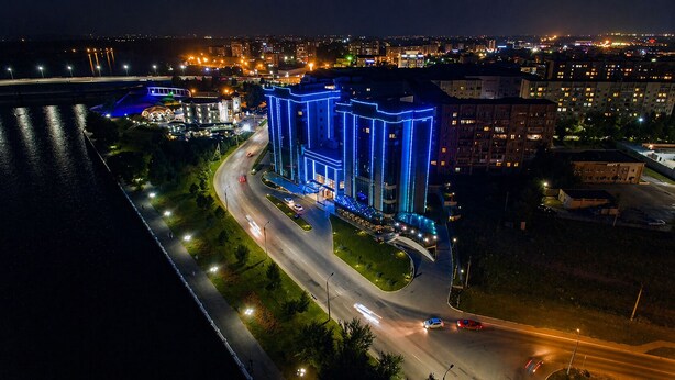 A modern building with a prominent entrance, adjacent to a river, with a well-lit roadway and surrounding greenery. - Hayat Hospitality Oskemen (Oskemen)