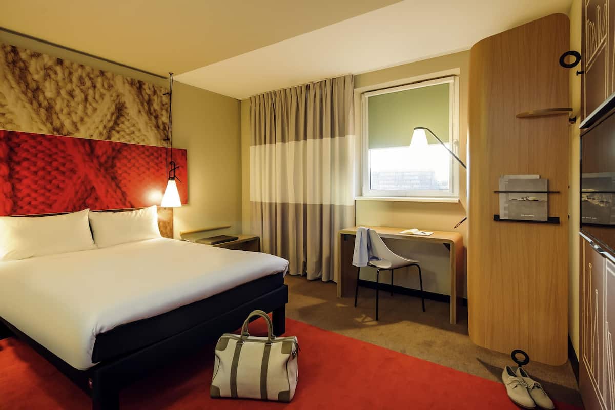 Una habitación de hotel con cama, escritorio, silla y ventana con cortinas.