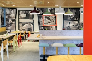 Una cafetería moderna con sillas de colores, una zona de bar central y fotografías blanco y negro en las paredes.