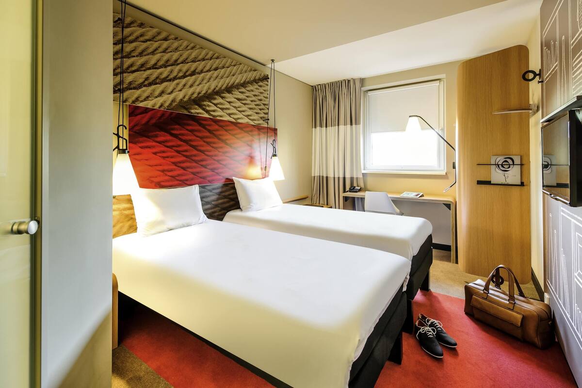 Een hotelkamer met twee bedden, een bureau, een stoel, een televisie, een klok en een handtas op de vloer.