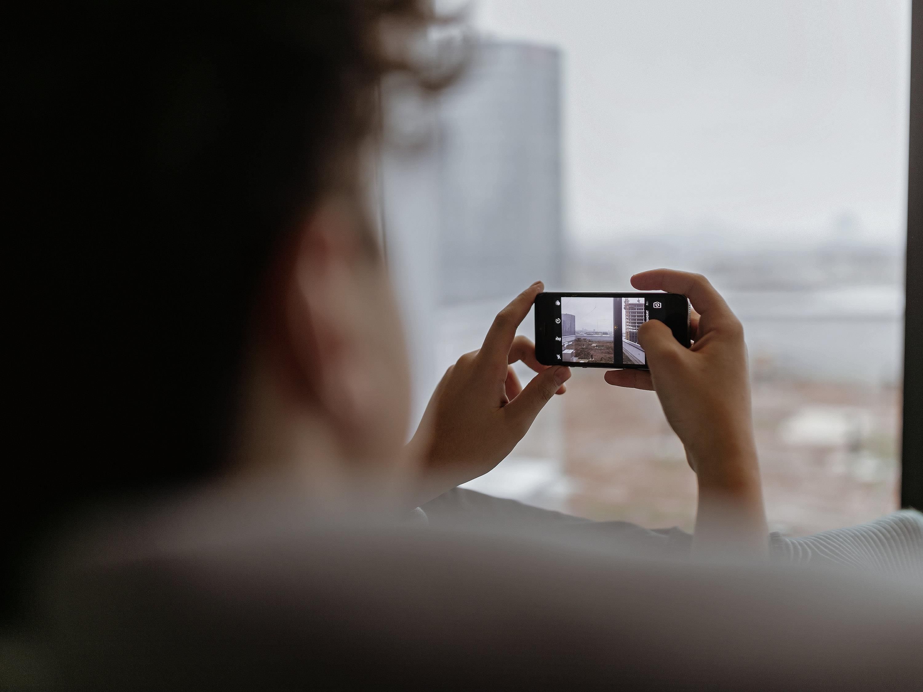 A person holding a smartphone, capturing a view through a window.