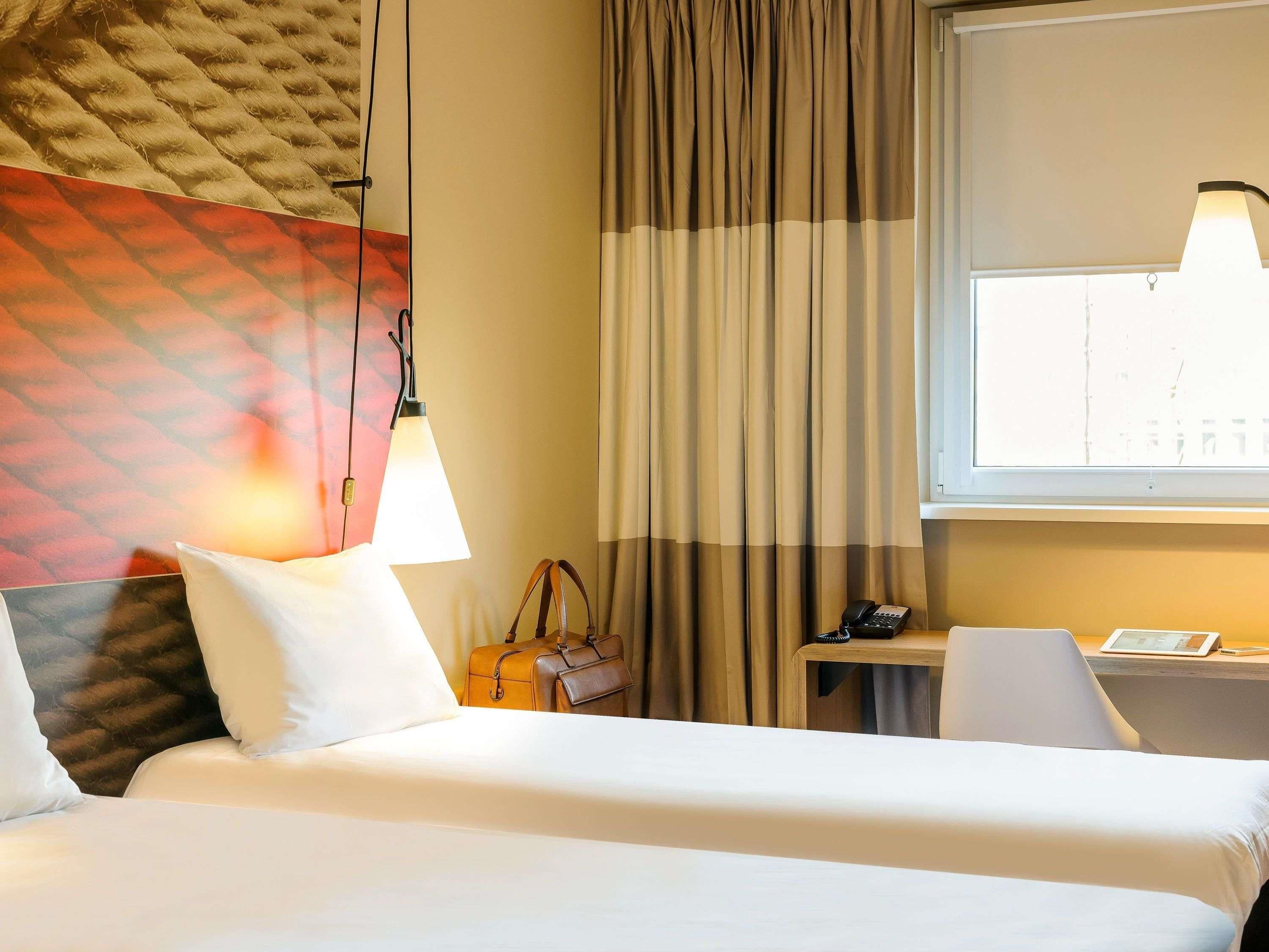 A hotel room with a bed, a desk, a chair, a lamp, a handbag, and a window with curtains.