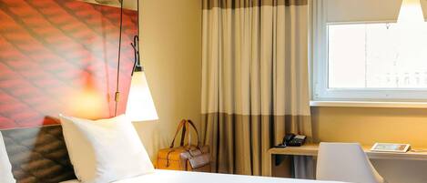 A hotel room with a bed, a desk, a chair, a lamp, a handbag, and a window with curtains.