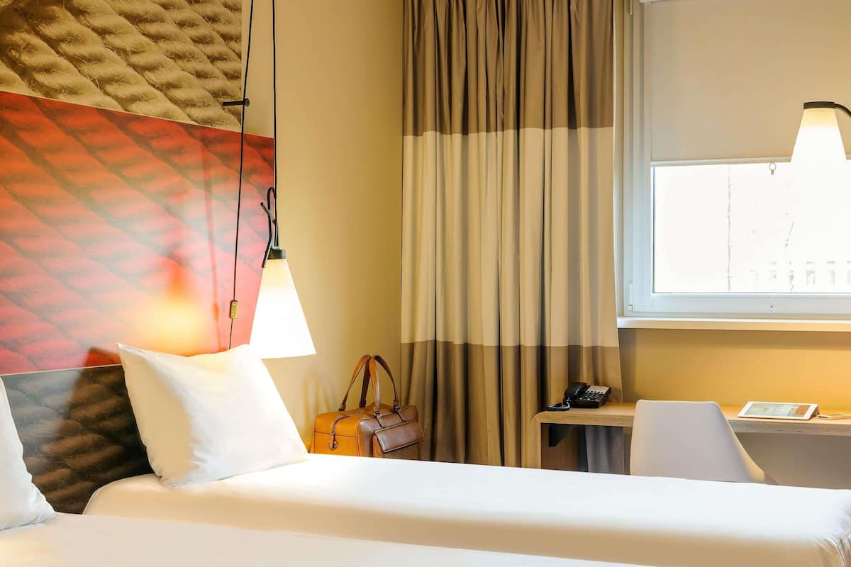 Une chambre d’hôtel avec un lit, un bureau, une chaise, une lampe, un sac à main et une fenêtre avec des rideaux.