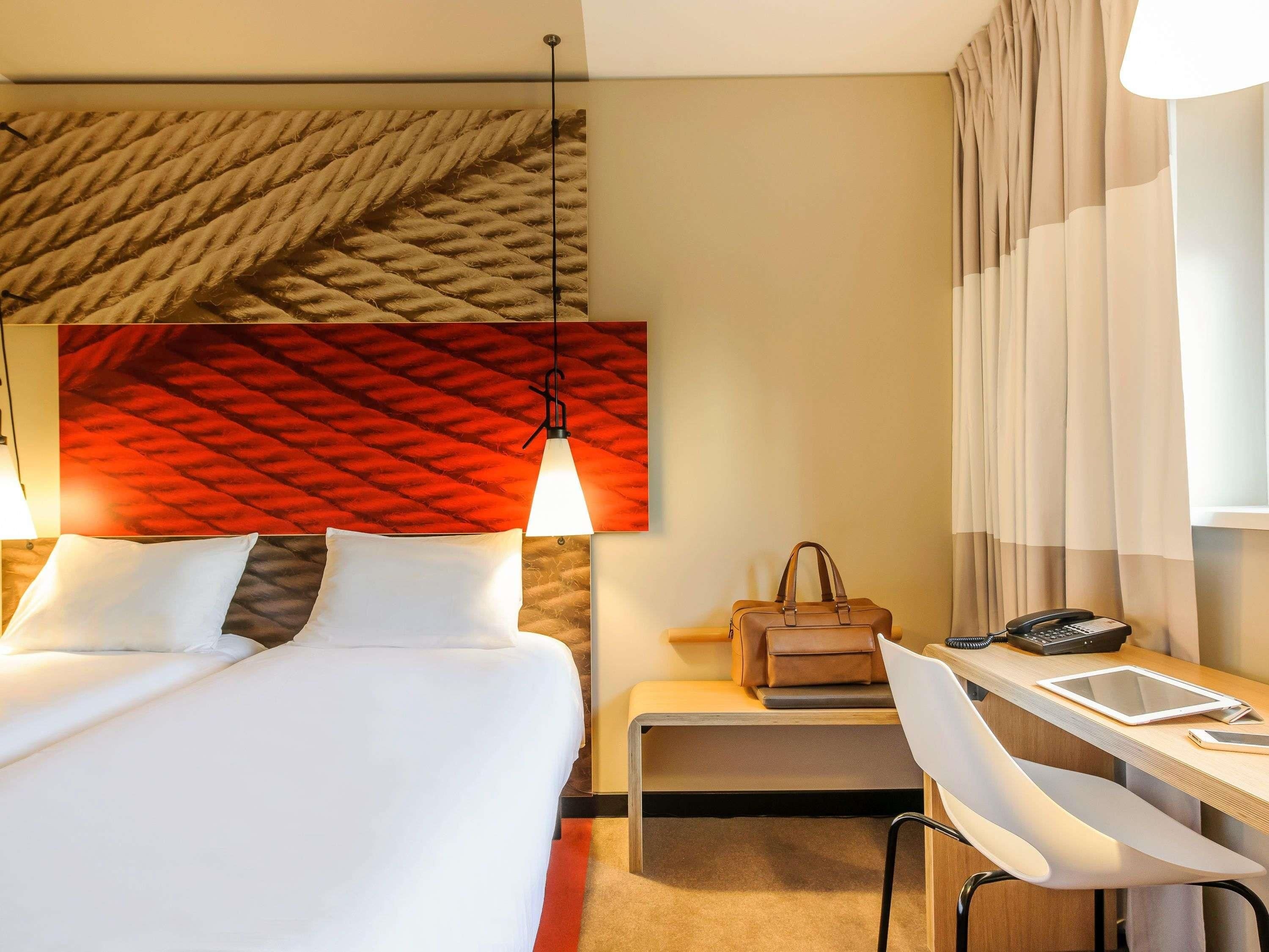 A hotel room with a bed, bedside lamps, a desk with a chair, a handbag, and a phone.