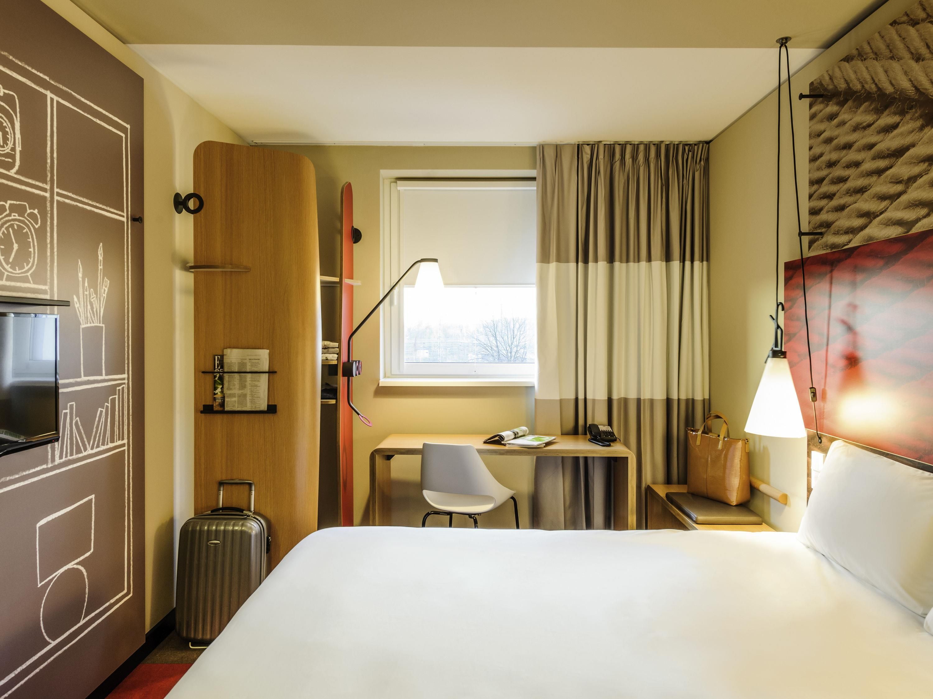 Habitación de hotel con cama, escritorio con silla, lámpara, maleta y armario.
