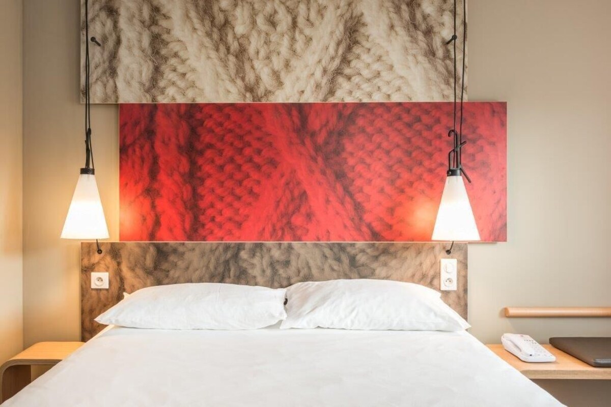 A hotel room with a bed, two bedside lamps, a nightstand with a phone, and a red abstract painting above the bed.