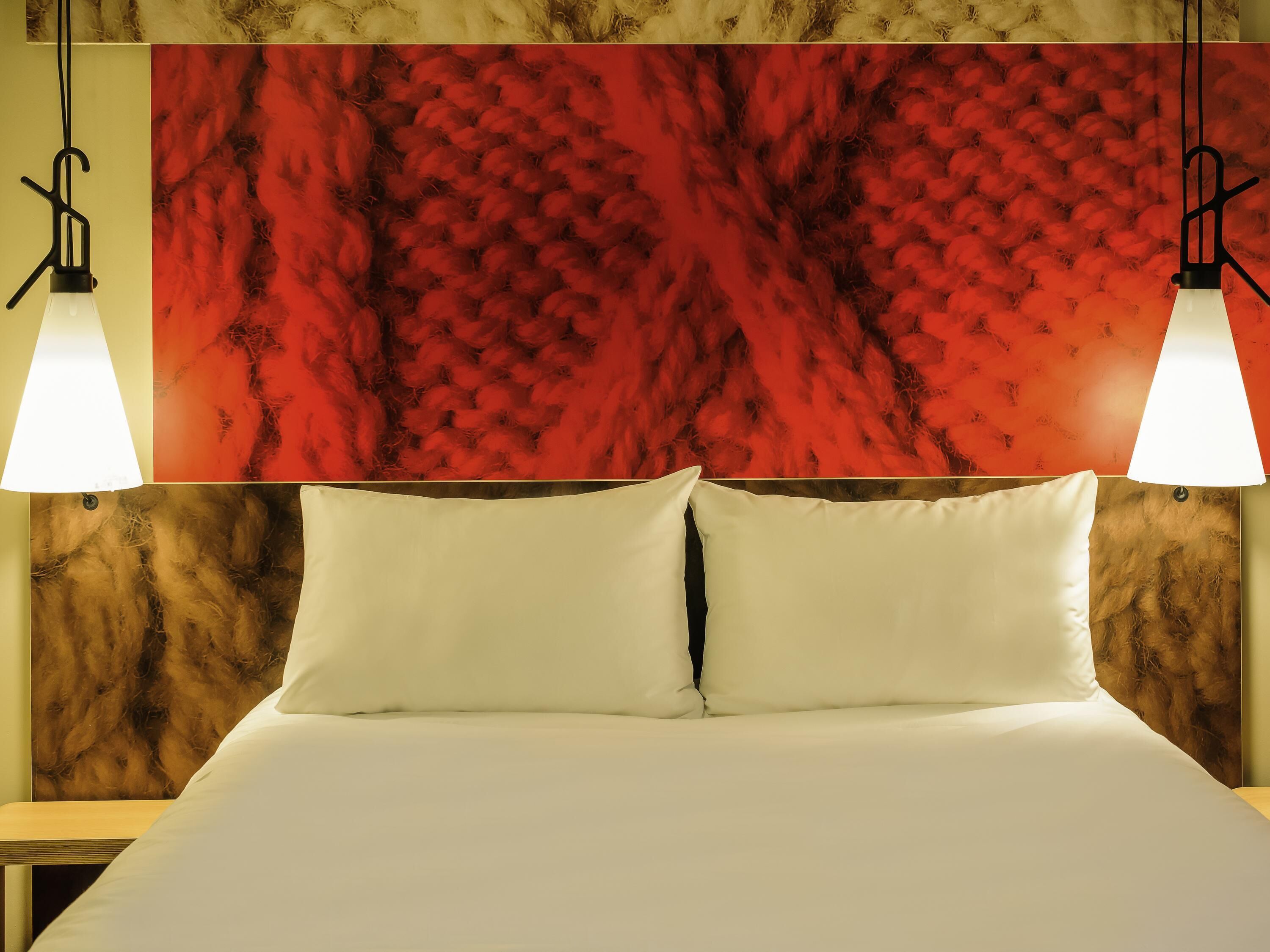 Una cama con ropa blanca, dos lámparas de noche y una pared roja con textura.