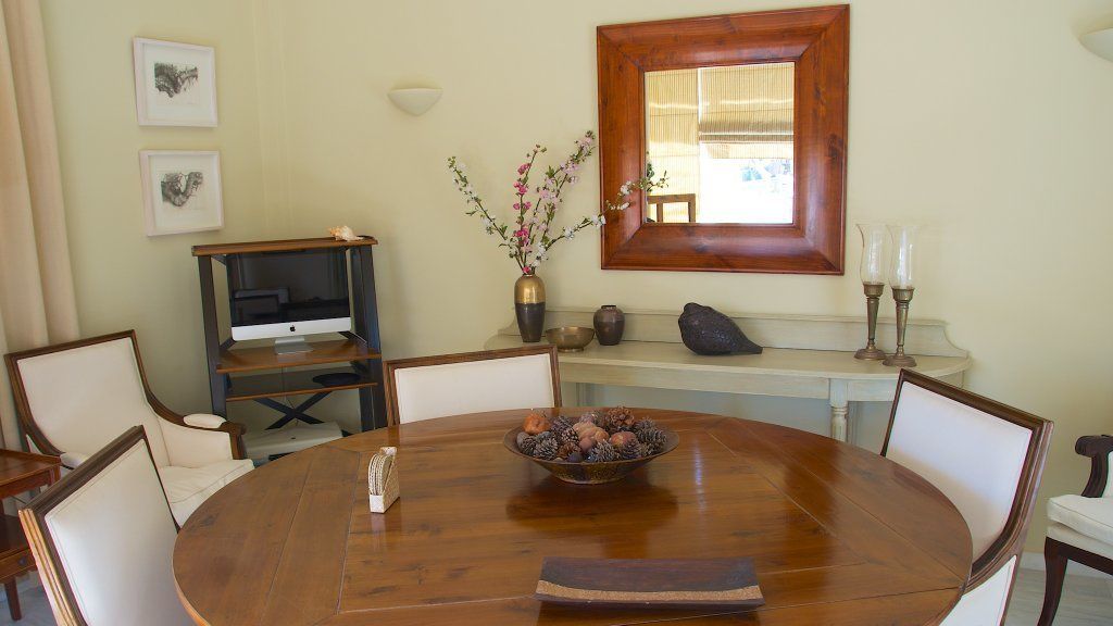 Un área de comedor con una mesa redonda de madera, sillas, un televisor sobre un mueble, un florero con flores y una ventana con marco de madera.