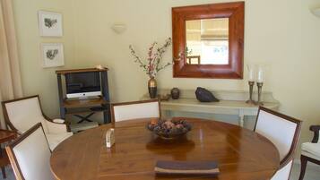 Un área de comedor con una mesa redonda de madera, sillas, un televisor sobre un mueble, un florero con flores y una ventana con marco de madera.