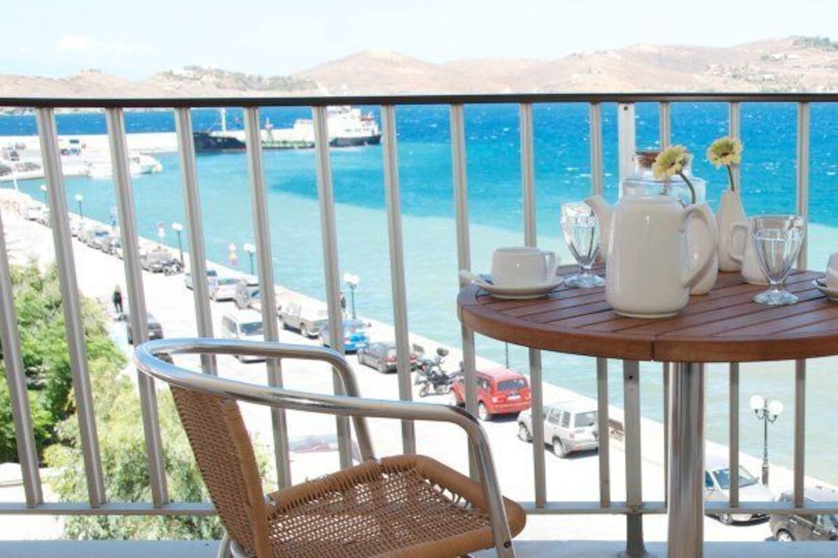 A balcony with a table set for two, overlooking a coastal area with a view of the sea and boats.