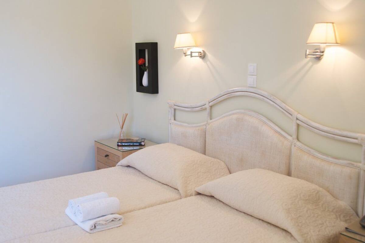 A neatly made bed with two pillows and a folded towel, a bedside table with a book and a vase of flowers, and two wall-mounted lamps.