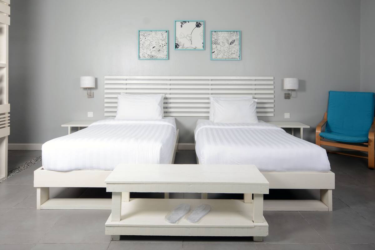 A double bed with white bedding, a small table with a pair of slippers, and a blue chair.