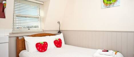 A neatly made bed with white bedding and two red strawberry pillows, a window with blinds, and a wall-mounted picture of a kiwi.