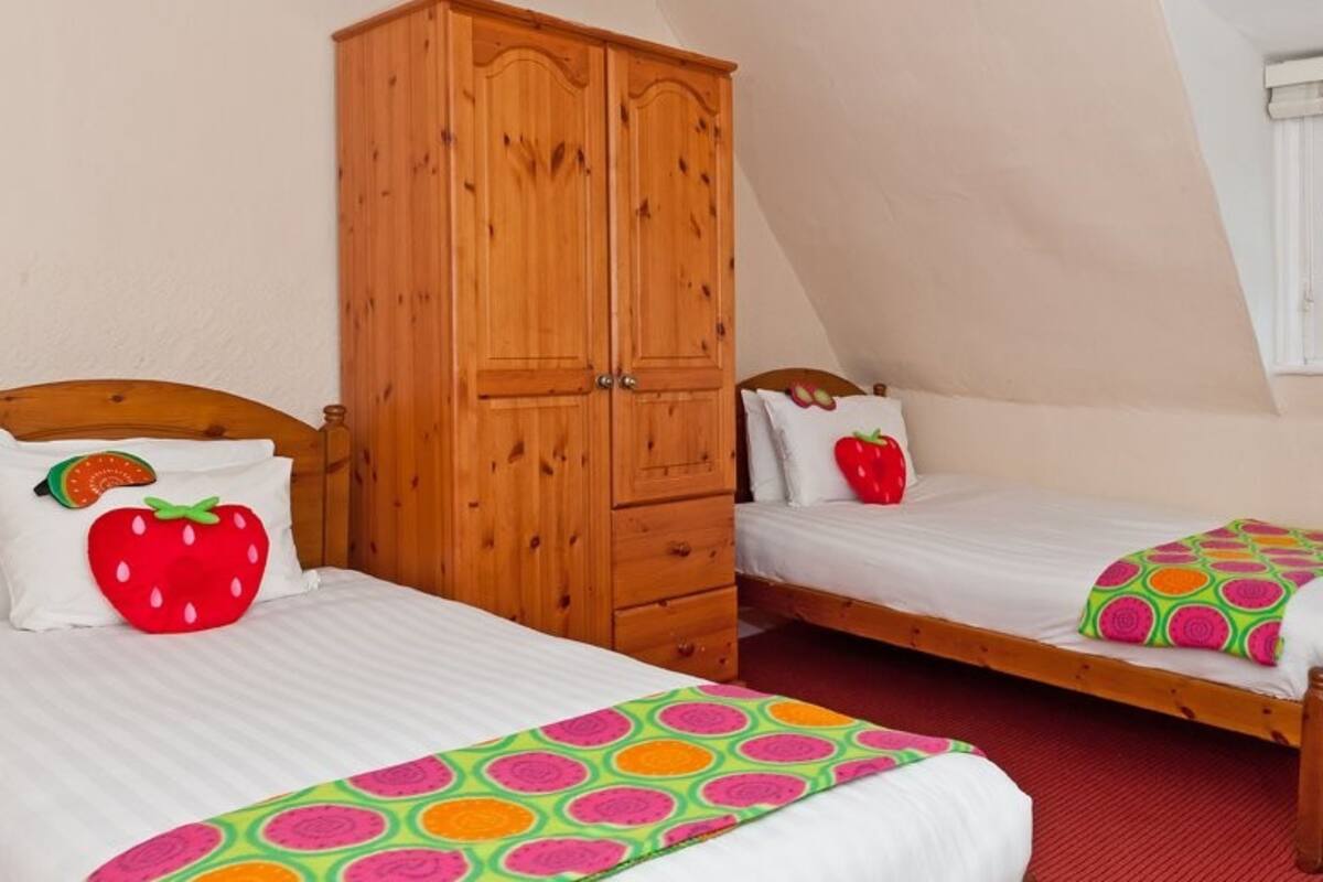 Un dormitorio con dos camas individuales, cada una con una colcha de colores y una almohada con motivos de fresas. Se observa un armario de madera, y la habitación tiene un techo inclinado.