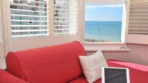 Un sofá rojo con una computadora portátil y un cojín con texto, una ventana que ofrece vistas al mar y a edificios, y una baranda blanca.