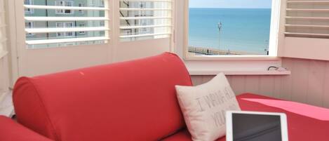 A red sofa with a laptop and a pillow with text, a window with a view of the sea and buildings, and a white railing.