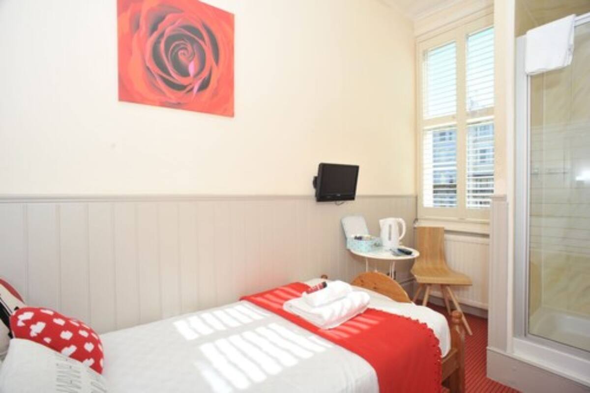 Una habitación de hotel con cama, colcha roja y blanca, un cuadro de una rosa roja en la pared, una mesita con una silla y una ventana con persianas.