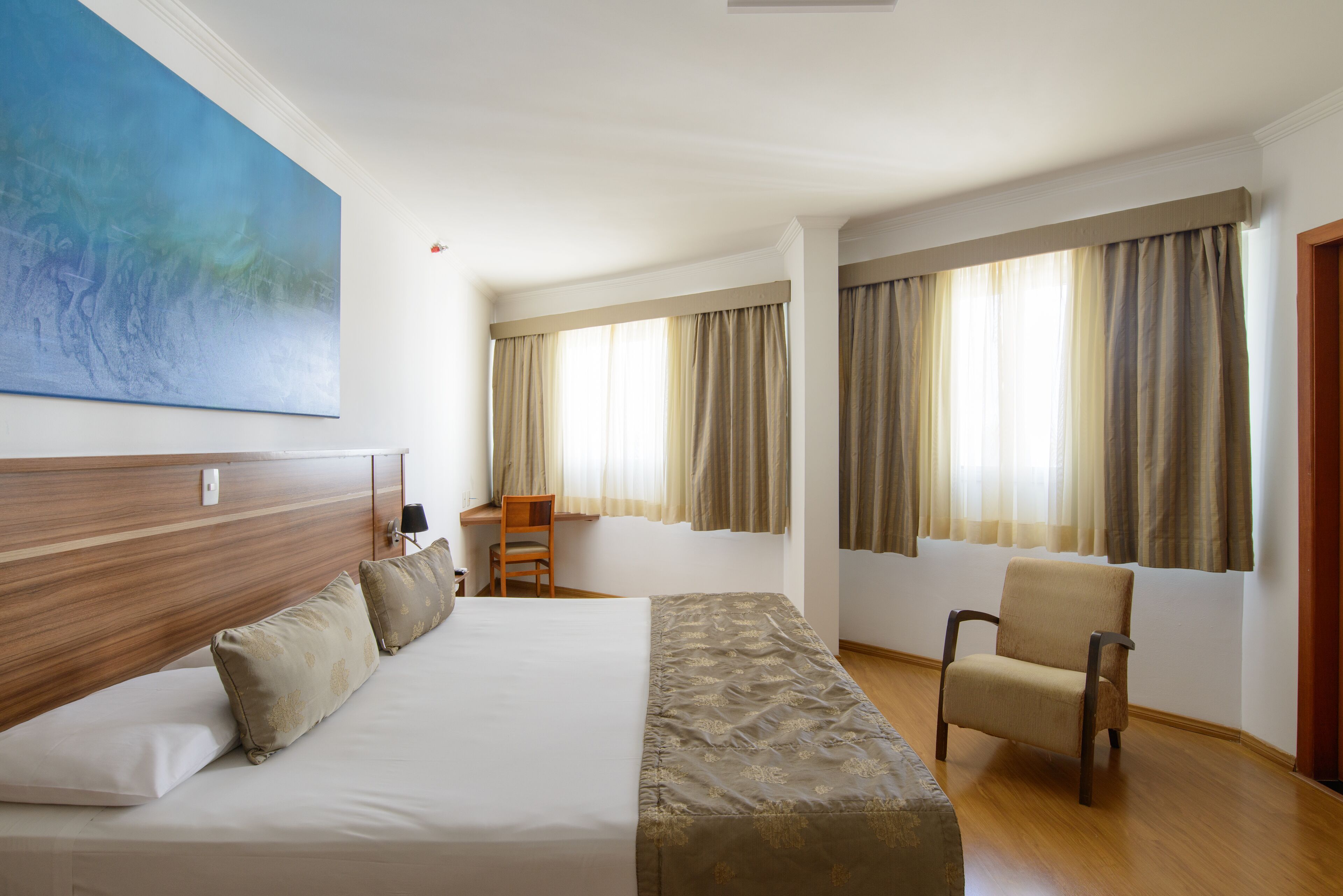 A hotel room with a large bed, a desk, a chair, and a painting on the wall.