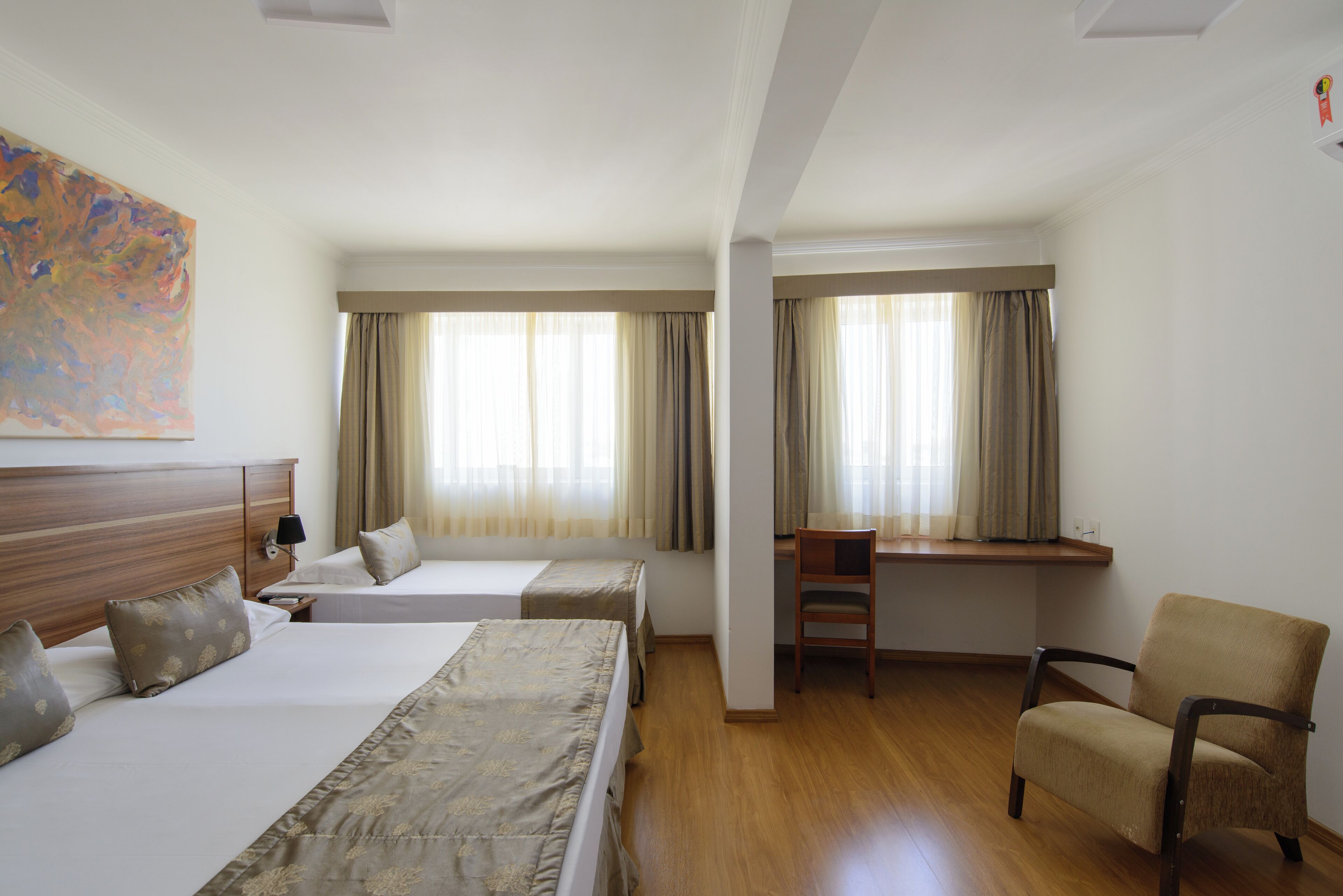 A hotel room with two beds, a desk, a chair, and large windows with curtains.