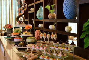 Un bufé con diversos postres y bebidas, una pared decorativa con jarrones, y una planta.