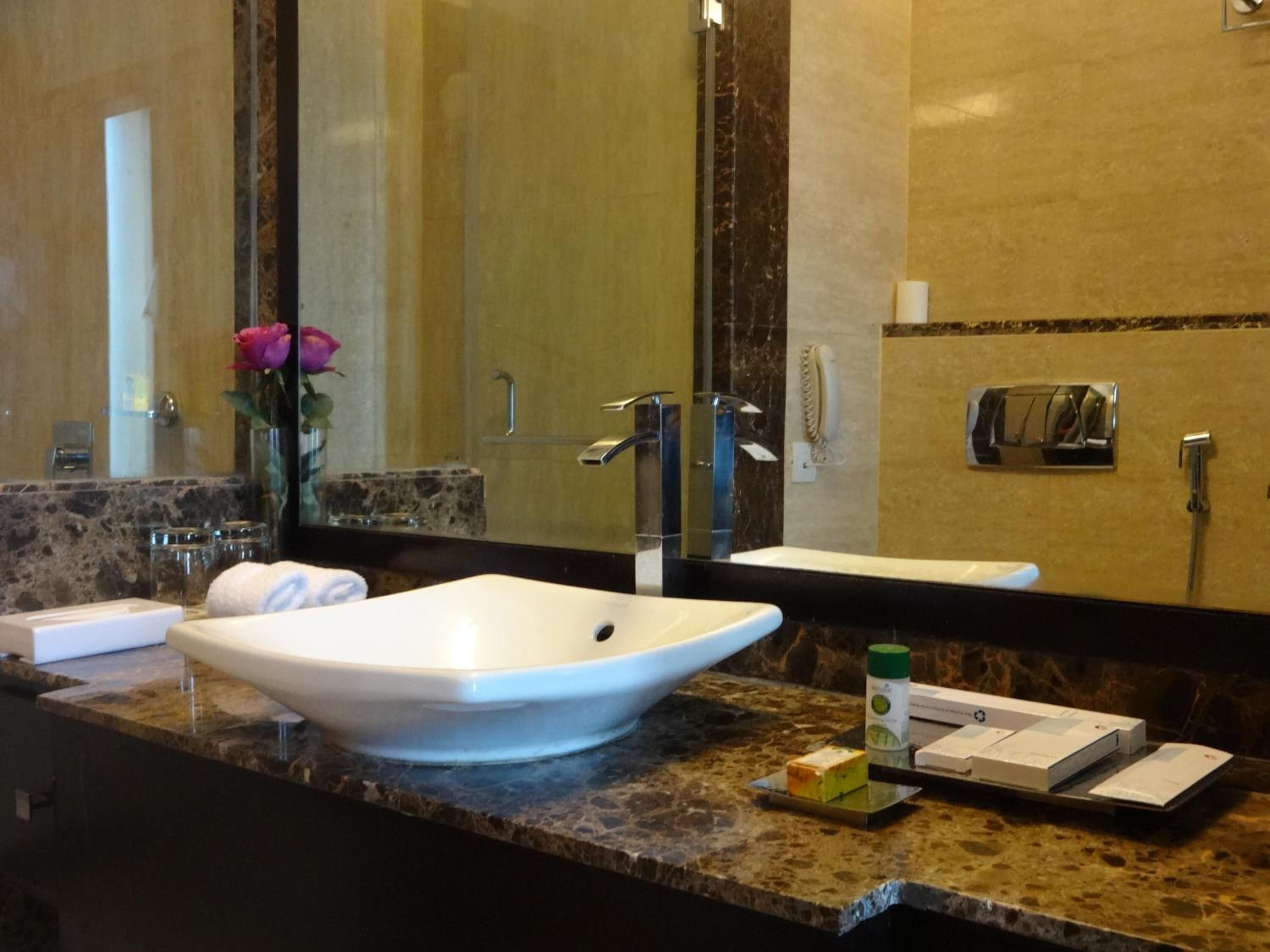 Un baño con un lavabo blanco tipo recipiente, un espejo grande, encimera de mármol y un florero con flores rosas.